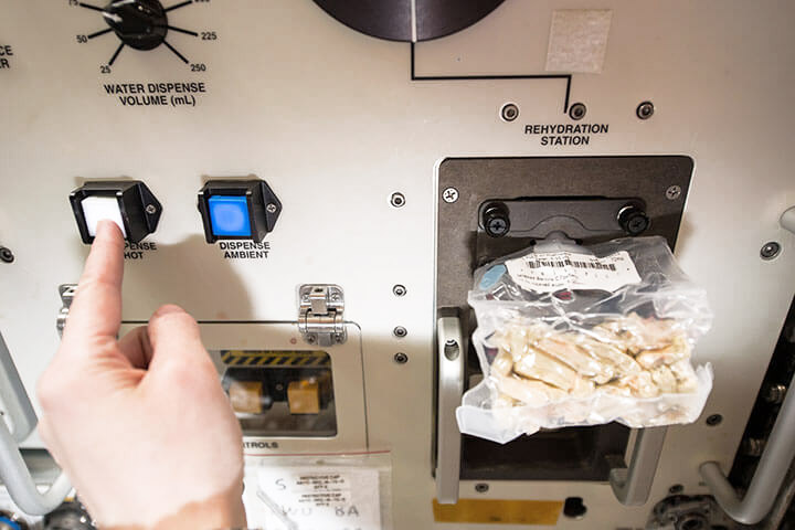 An astronaut prepares her bagged lunch using a space food rehydrator aboard the International Space Station (ISS).