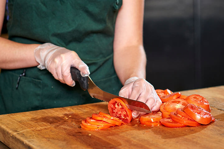 Tomatoes are cut on their own cutting board, with gloves and a clean knife. 