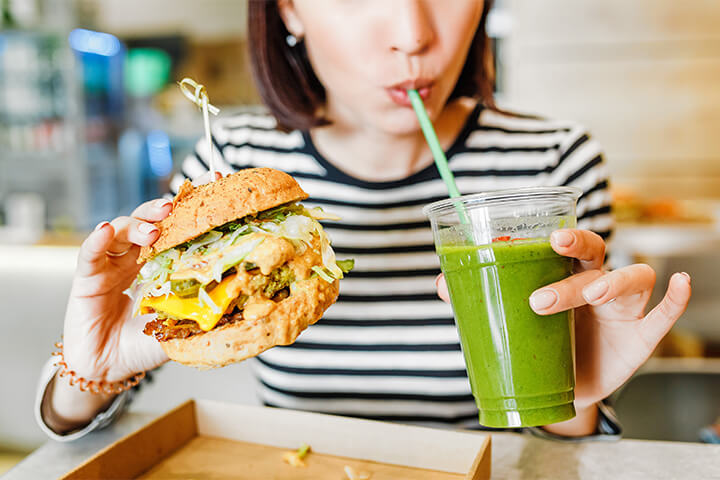 A woman holds an extravagant burger and sips green juice.