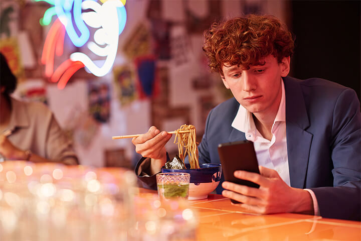 A sad boy looks at his phone while he eats asian noodles of some sort. Would a phone ban help?