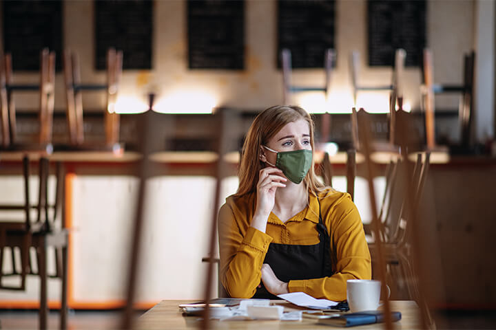 A sad masked employee sits alone in an empty restaurant, thinking of days gone by.